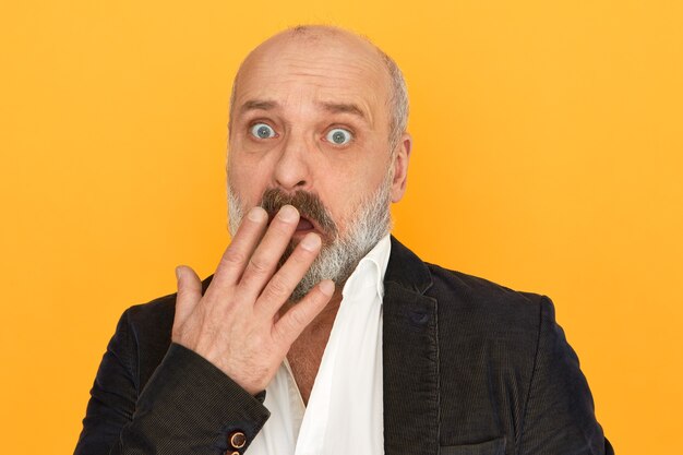 Funny bug eyed surprised male pensioner with thick beard gasping covering mouth with hand, receiving bad unexpected news, having fearful puzzled facial expression