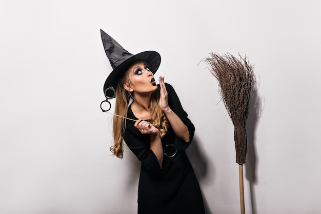 Foto gratuita divertente ragazza caucasica in posa in costume da strega nel carnevale. donna dai capelli lunghi in cappello magico in piedi sul muro bianco con la scopa.