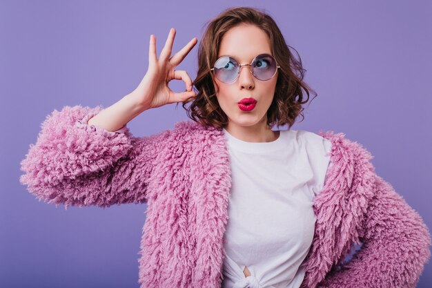 Funny caucasian woman in glamorous fur coat posing . Cheerful female model with wavy brown hair making faces during photoshoot.