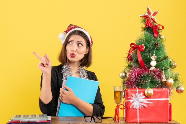 Funny charming lady in suit with santa claus hat and new year decorations holding document thinking deeply in the office on yellow isolated 