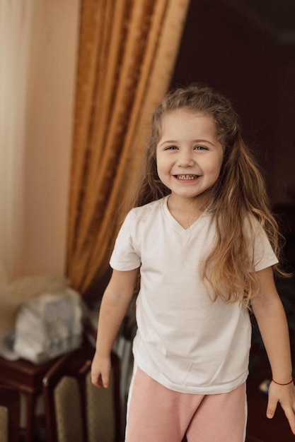 La bambina affascinante e divertente con lunghi capelli ondulati vestita con una maglietta bianca si diverte a casa ballando e sorridendo