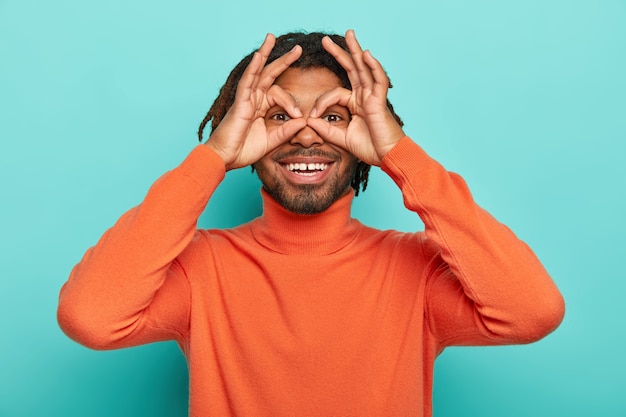 Funny cheerful guy holds hands near eyes, pretends looking through binoculars, has dreadlocks wears orange turtleneck isolated on blue