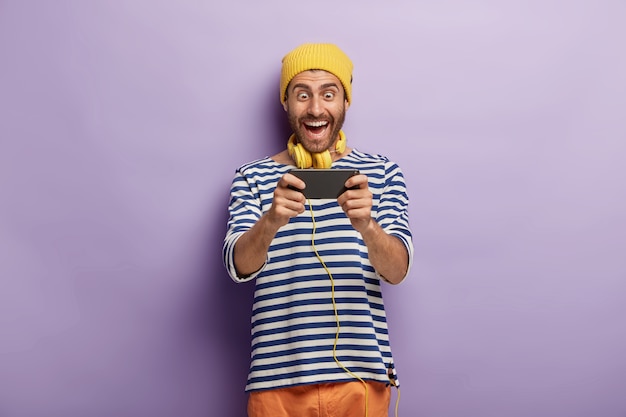 Funny cheerful male gamer plays video games via smartphone, wears yellow hat and striped jumper, being addicted to modern technologies, isolated on purple wall, checks out new application