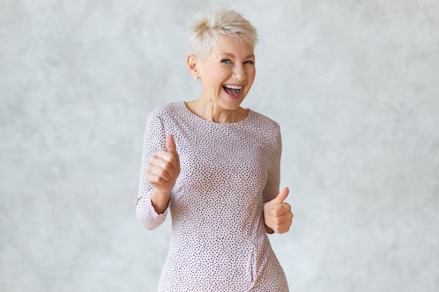 Funny cheerful middle aged blonde woman wearing elegant pencil dress dancing and showing thumbs up gesture as sign of approval, celebrating success or profitable deal, smiling broadly