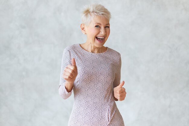 Funny cheerful middle aged blonde woman wearing elegant pencil dress dancing and showing thumbs up gesture as sign of approval, celebrating success or profitable deal, smiling broadly