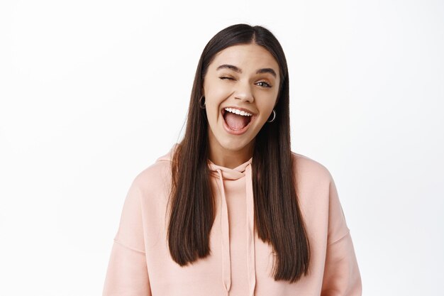 Funny and cheerful young beautiful girl, winking and showing tongue, having fun, standing over white wall