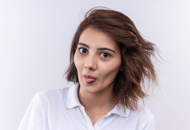 Funny cheerful young girl with short hair wearing white polo shirt looking at camera sticking out tongue 