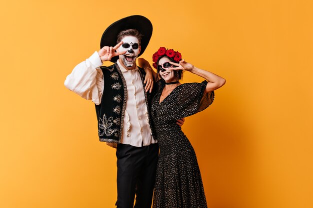 Funny couple in great mood posing on orange wall. Portrait of man and woman with Mexican-style face art showing peace sign.