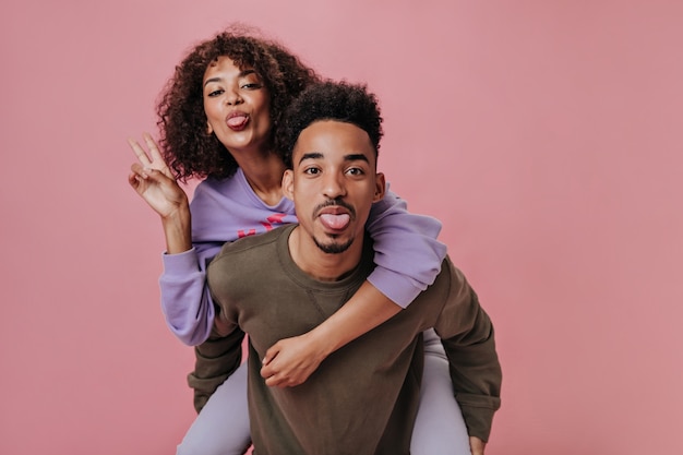 Funny couple in love showing tongues. Girl in purple outfit showing peace sign on pink wall