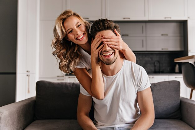 Funny couple playing seek and hide. Curly girl sitting on sofa with boyfriend.