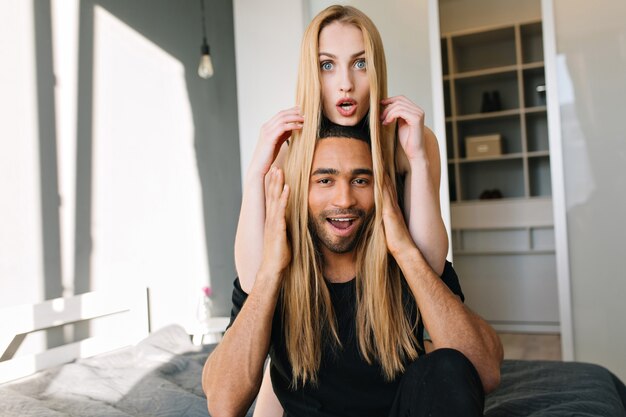 Funny couple of pretty young woman with long blonde hair and joyful handsome guy having fun on bed in modern apartment. Joy, happiness, morning together, love, leisure, home