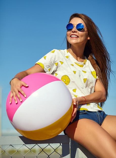 Funny crazy glamor stylish smiling beautiful young woman model in bright hipster summer casual clothes posing in the street behind blue sky and sitting on the fence. Playing with colorful inflatable b