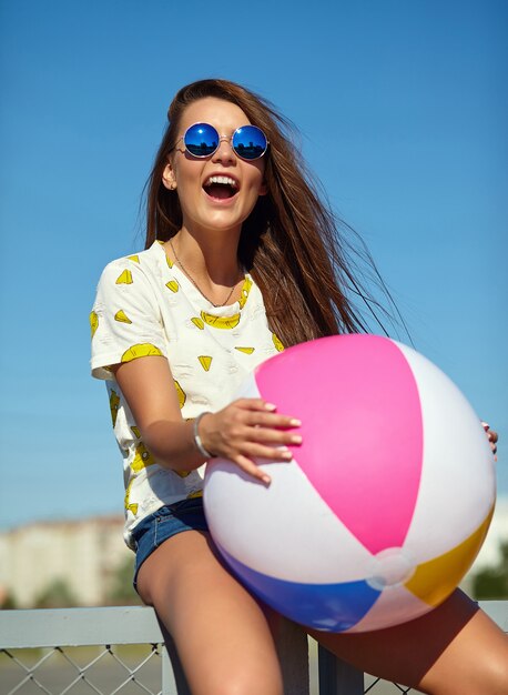 Funny crazy glamor stylish smiling beautiful young woman model in bright hipster summer casual clothes posing in the street behind blue sky and sitting on the fence. Playing with colorful inflatable b