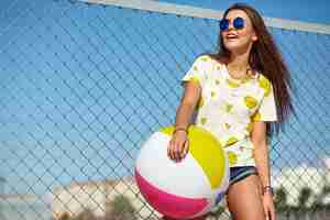 Free photo funny crazy glamor stylish smiling beautiful young woman model in bright hipster summer casual clothes posing in the street behind iron grating and blue sky. playing with colorful inflatable ball floa