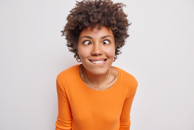 Funny crazy woman with curly hair crosses eyes bites lips foolishes around tries to amuse someone wears orange jumper isolated over white background grimaces at camera. Human face expressions