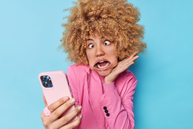 Free photo funny curly haired woman goes crazy crosses eyes uses mouth expander has white even teeth makes grimace wears formal pink jacket takes selfie via modern smartphone isolated over blue background