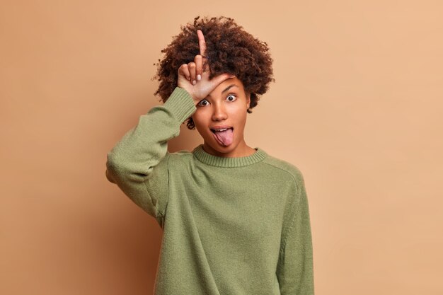 Funny curly haired young woman makes loser gesture sticks out tongue dressed in casual jumper isolated over brown wall mocks at someone who lost bet