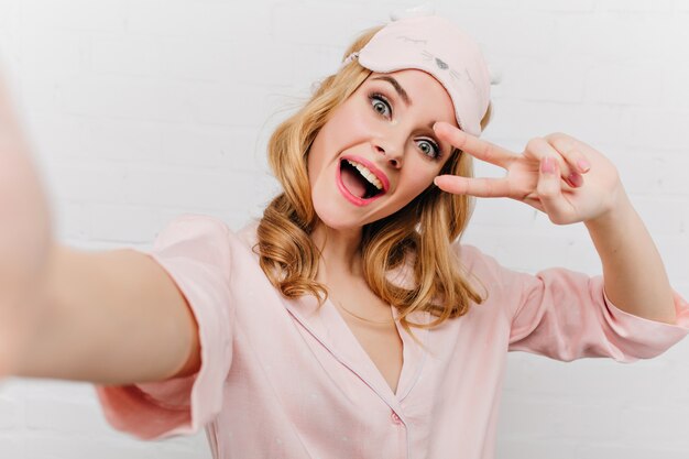 핑크 수면 마스크 만들기 selfie에 재미있는 곱슬 여자. 흰 벽에 고립 된 실크 잠옷에 매력적인 금발 소녀의 실내 자화상.
