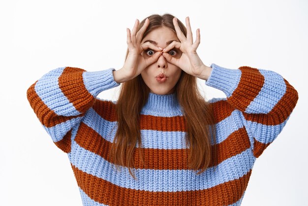 Funny cute girl look through hand binoculars pucker lips saying wow making glasses gesture around eyes and staring at camera standing in sweater against white background