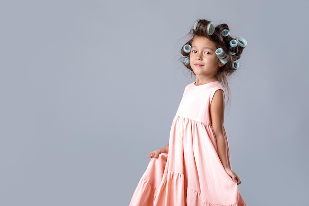 Funny cute little child girl in pink dress and hair curlers posing on gray background. human emotions and facial expression
