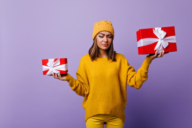 Funny dark-haired lady in stylish hat holding present boxes. Portrait of appealing brunette woman in yellow sweater posing before christmas.