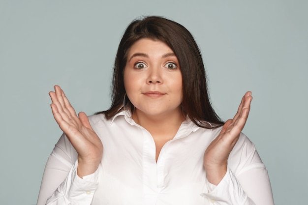 Funny dark haired overweight female throwing out her hands, being at loss, having confused puzzled look, her eyes full of surprise and astonishment