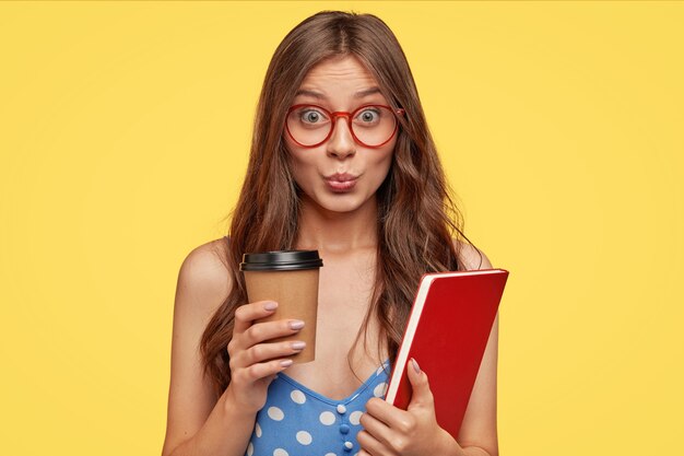 Funny dark haired young European girlfriend keeps lips pressed together, wears transparent glasses, holds coffee in disposable cup