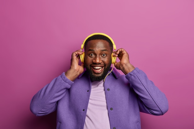 Free photo funny dark skinned bearded man listens music in headphones from some modern device, keeps hands on ears, enjoys good sound, has good mood