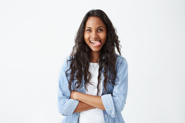 Funny dark-skinned female with loose black hair and charming dark eyes having joy while showing tongue, keeping her arms folded. Cheerful female student wearing denim shirt having fun after classes lo