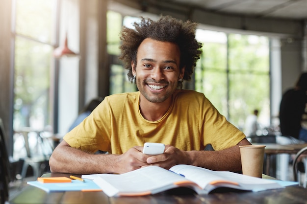 Uomo dalla carnagione scura divertente con l'acconciatura africana che lavora sulla carta del corso mentre era seduto in un caffè durante la pausa pranzo che tiene lo smartphone felice di finire il suo lavoro. ragazzo africano con un ampio sorriso nella caffetteria