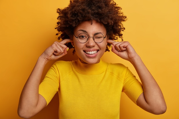 Funny dark skinned woman plugs fingers in ears, cannot concentrate because of noise in crowded place, winks eye and shows white teeth, wears spectacles and tshirt, isolated on yellow wall