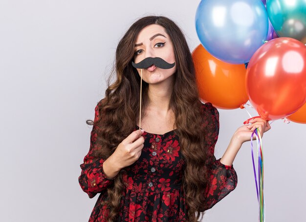 Funny doubtful young party girl holding balloons and fake mustache on stick above lips looking at camera with pursed lips isolated on white wall