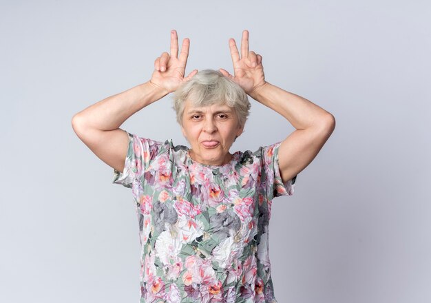 Funny elderly woman puts hands on head gesturing horns and sticks out tongue isolated on white wall