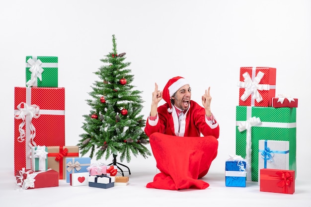 Free photo funny emotional excited young man dressed as santa claus with gifts and decorated christmas tree sitting on the ground on white background