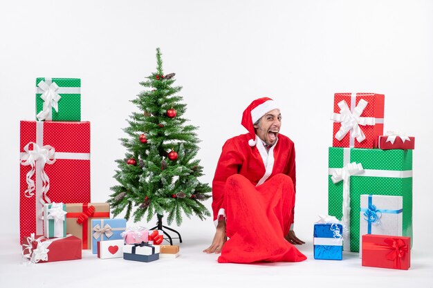 Funny emotional young man dressed as Santa claus with gifts and decorated Christmas tree sitting on the ground on white background