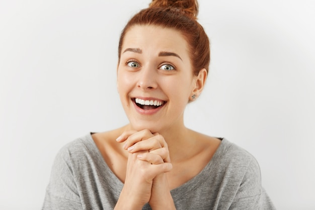 Free photo funny emotional young redhead woman with hairbun keeping hands clasped as if trying to be nice, flattering someone, asking for something. human facial expressions and emotions. body language