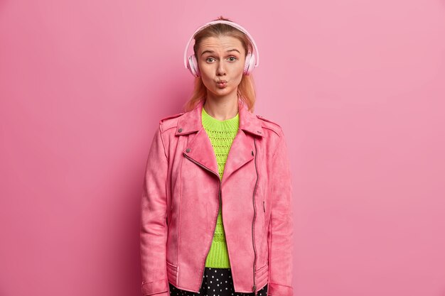 Funny European girl keeps lips rounded, listens music in headphones, picks song in playlist, being on her way to school, dressed in fasionable pink jacket, enjoys favorite singer