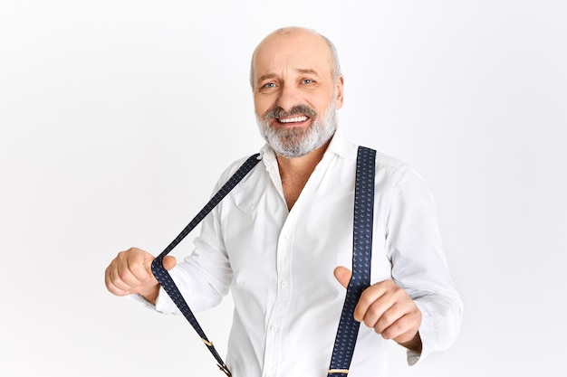 Funny European male pensioner with bald head and gray beard posing isolated wearing elegant white shirt, adjusting suspenders, going to dinner