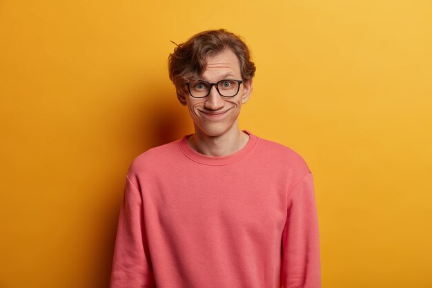 Funny European man has pleased expression, smiles joyfully, wears optical glasses and rosy jumper, hears good news, isolated over yellow wall, express good emotions. Male nerd in spectacles