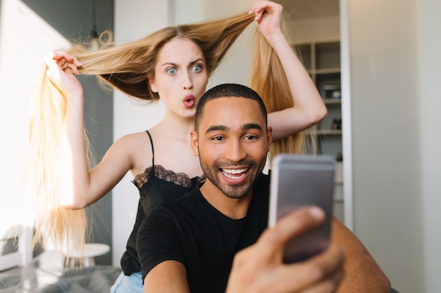 Funny excited young woman having fun with her long blonde hair behind smiled handsome guy making selfie of them on bed in modern apartment. Lovers, happiness, entertainment