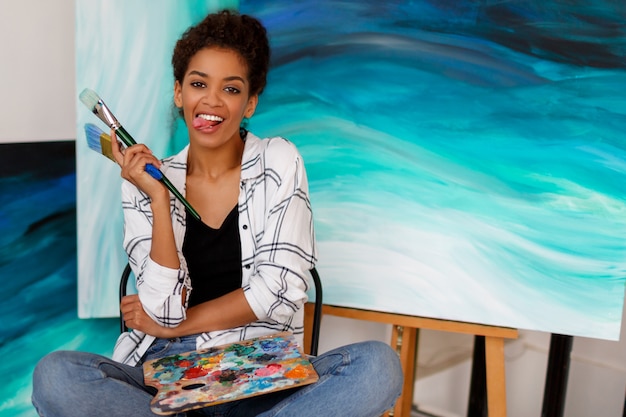Free photo funny female artist  sitting with amazing abstract sea acrylic hand drawn artwork at the studio. holding brushes and palette.