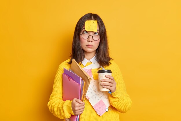 Funny female nerdy student crosses eyes has sticky note stuck on forehead has coffee break while preparing for exam much work to do holds folders and papers.