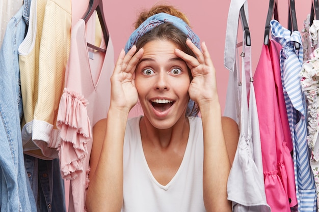 Free photo funny female seller of clothes looking into distance waiting for buyers while standing near rack with clothes. happy woman keeping hands on forehead looking into distance waiting for someone