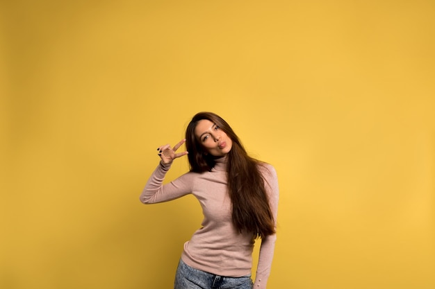 청바지와 분홍색 셔츠를 입고 긴 갈색 머리를 가진 재미있는 친절한 소녀는 키스를 보내고 노란색 벽 위에 평화 기호를 표시합니다.