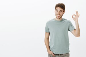 Funny friendly-looking european male coworker in trendy t-shirt, raising hand in okay or fine gesture and smiling broadly