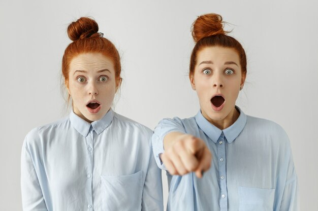 Funny frightened sisters wearing identical hair knot and light-blue shirts having shocked looks, scared with horror movie they watching together at home, one of girls pointing finger