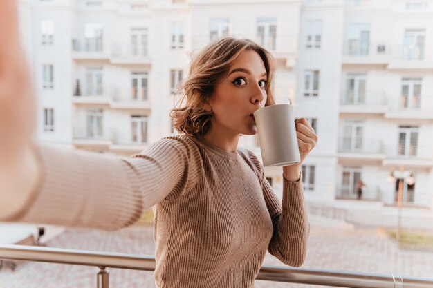 バルコニーでコーヒーを飲む茶色のセーターの面白い女の子。お茶で自分撮りをする魅力的な若い女性。