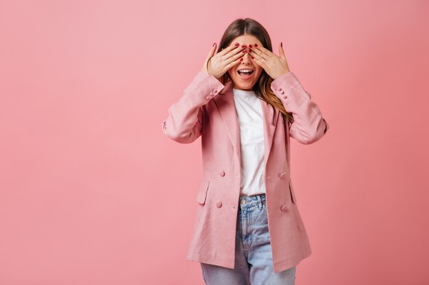 目を覆うカジュアルなジャケットの面白い女の子。ピンクの背景に分離されたトレンディな女性のスタジオショット。