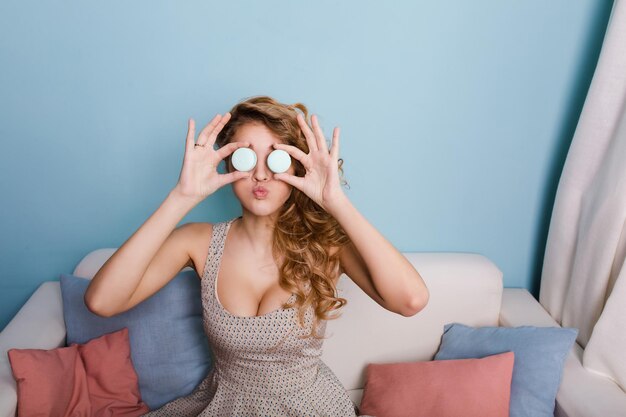 Funny girl making faces with two macaroons placed instead of eyes. Girl has curly blond hair and looks sexy. She sits on white sofa with colorful pillows in a studio with white walls.