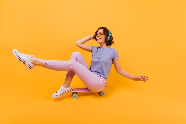 スケートボードに座って顔を作るピンクのズボンの面白い女の子。ヘッドフォンで素敵な白人女性の屋内写真